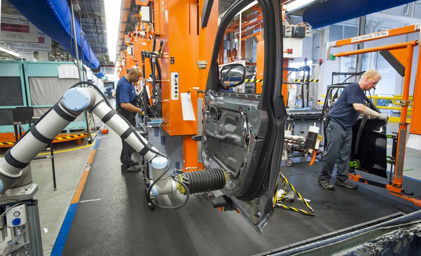 A humanoid robot handling a sheet metal part on a car assembly line next to a human worker.